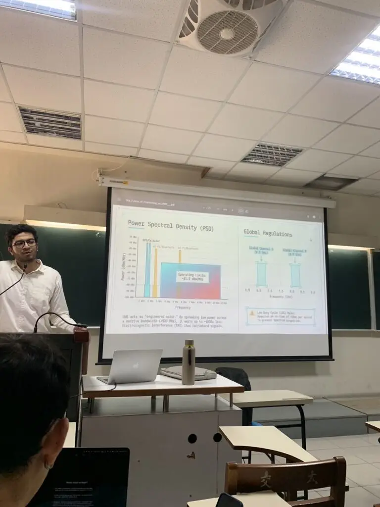 A man presenting a slideshow on Power Spectral Density and global regulations during a classroom lecture.