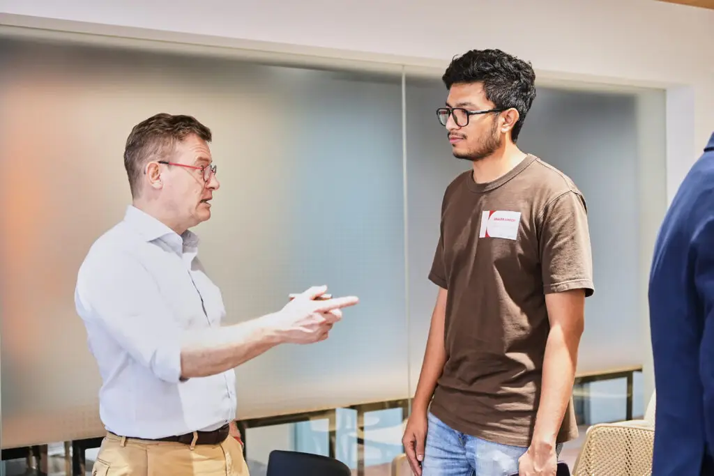 Older businessman gesturing while talking to a young professional at a corporate networking event.